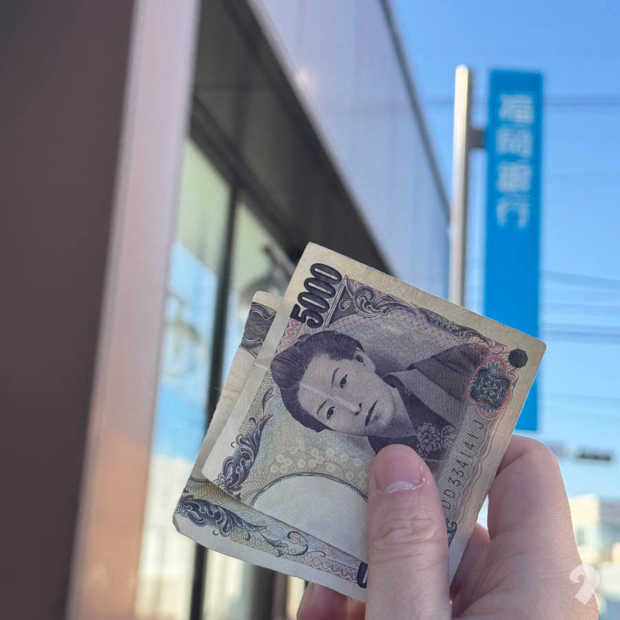 在福岡銀行藍色戶外招牌前，手中拿著撕成兩半的 5,000 日圓鈔票。