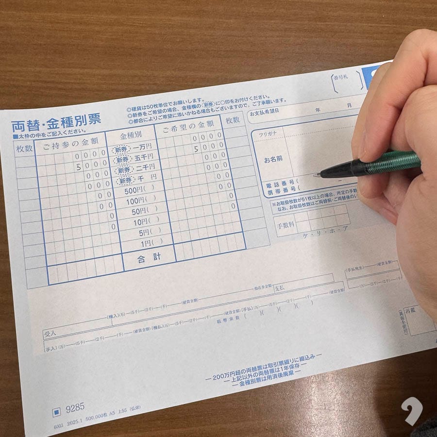 手持原子筆在木製櫃檯上填寫日文「兩替・金種別票」（貨幣兌換/幣值指定表單）的特寫。