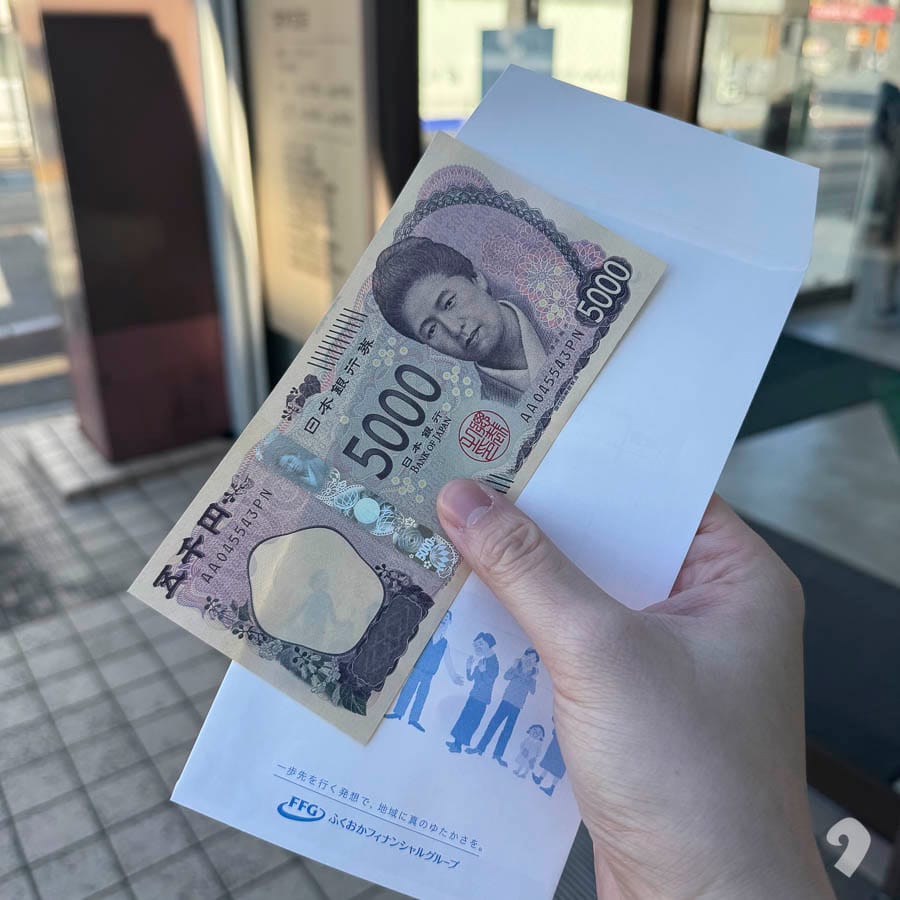 手中拿著一張新款日幣 5,000 元鈔票，下方墊著福岡金融集團（FFG）的白色信封。