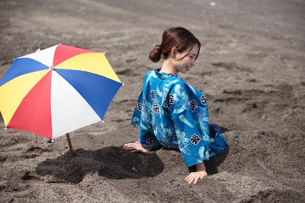 A woman in a blue yukata settling into the black volcanic sand at an Ibusuki sand bath, next to a colorful beach umbrella