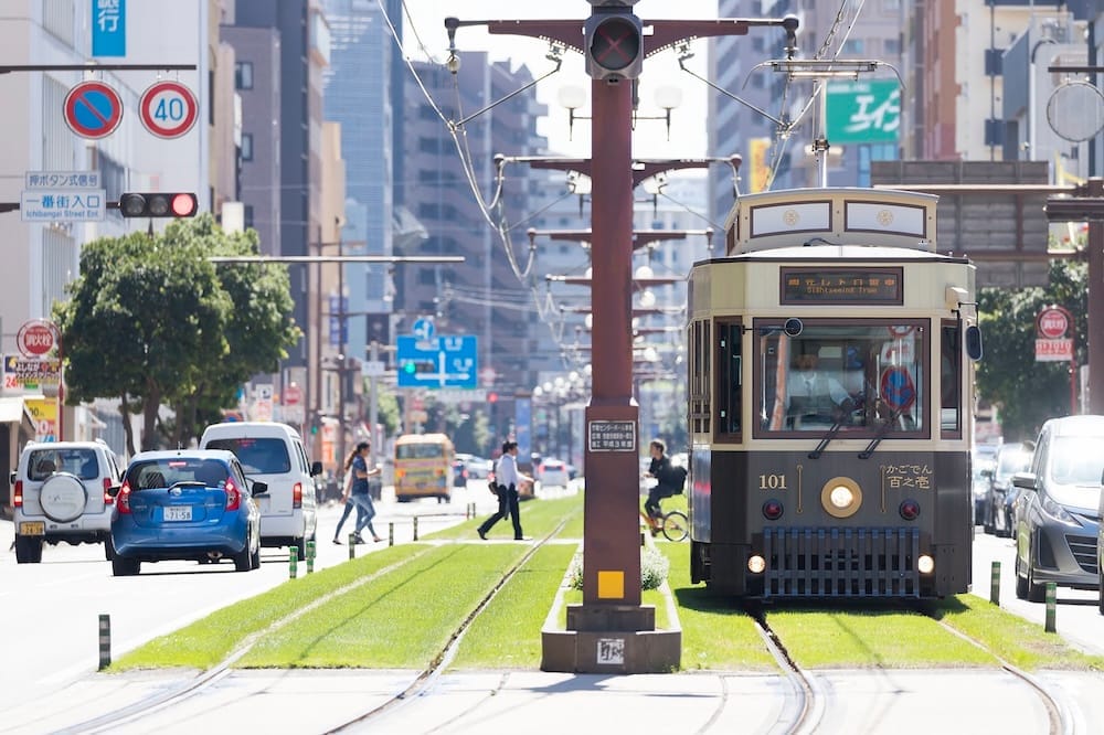 A vintage-style Kagoshima City streetcar running on grass-covered tram tracks, with office buildings and pedestrians on either side