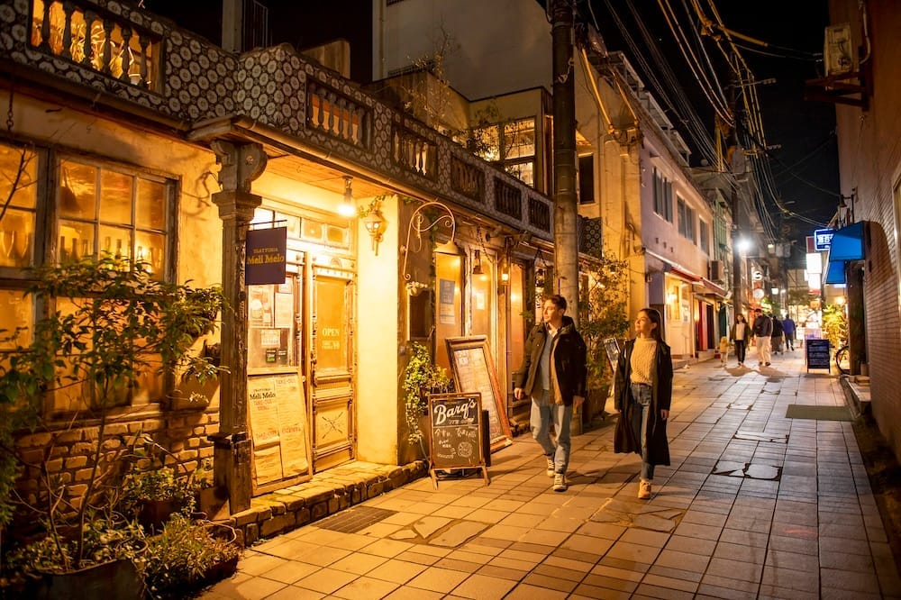 A couple strolling past warmly lit, retro-style storefronts on a stone-paved street at night