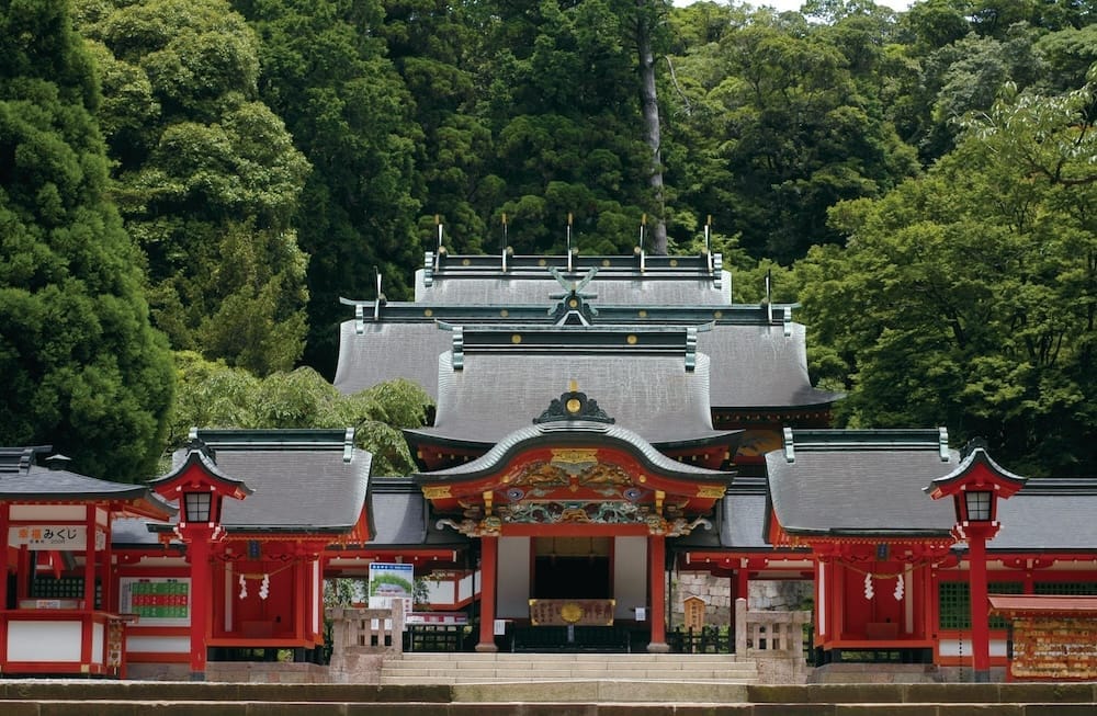 The ornate vermilion main hall of Kirishima Shrine, with layered gray roofs set against a thick green forest