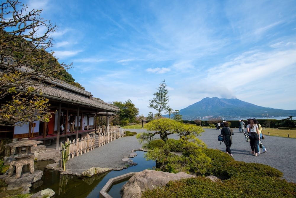 Pine trees and a garden pond at Sengan-en beside a wooden corridor, with Sakurajima rising in the distance under a blue sky