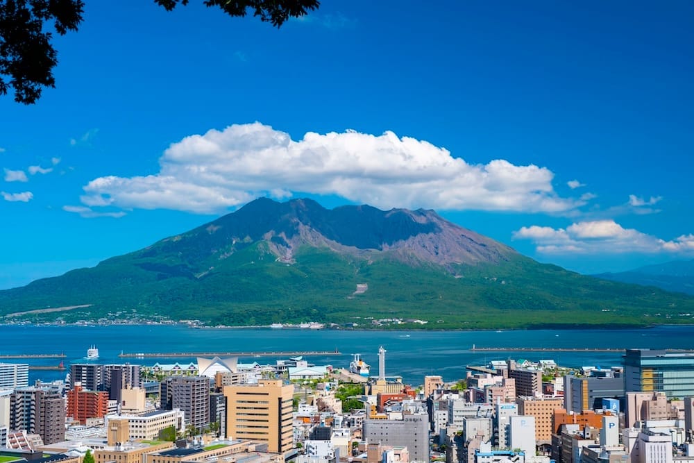 Sakurajima volcano under clouds on a sunny day, looking across Kinko Bay to the buildings of downtown Kagoshima in the foreground